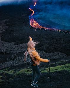 a woman standing on top of a lush green hillside next to a lava covered mountain