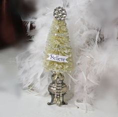 a small white christmas tree with a name tag on it's top and feathers around the base