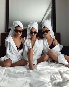 three women in white robes sitting on a bed with their feet up and one wearing sunglasses