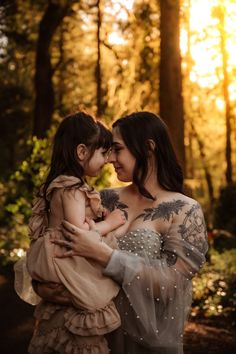 a woman holding a baby in her arms while standing in the woods with trees behind her