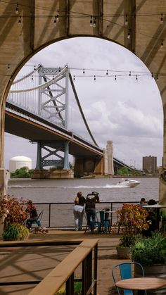 some people are standing under an overpass and looking at the water below them with a bridge in the background