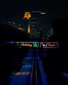 a train traveling down tracks next to tall buildings in the background at night with lights on