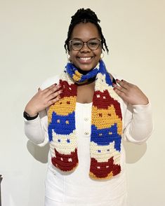 a woman wearing glasses and a crocheted scarf with cats on it, standing in front of a white wall