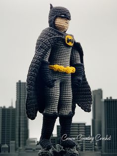 a crocheted batman figure standing in front of a cityscape