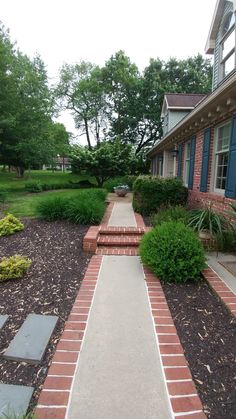 a brick walkway leading to a house with blue shutters and trees in the background