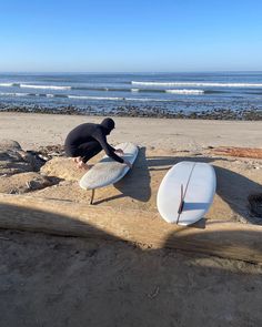 a man kneeling down next to two surfboards on top of a sandy beach near the ocean