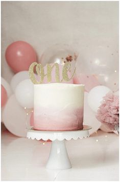 a pink and white ombreed cake with the word one on top sitting on a pedestal in front of balloons