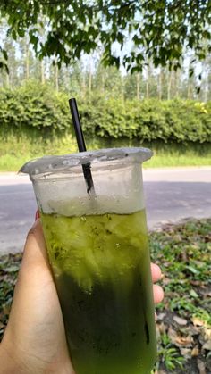 a person holding up a green drink in front of some bushes and trees with a black straw sticking out of it