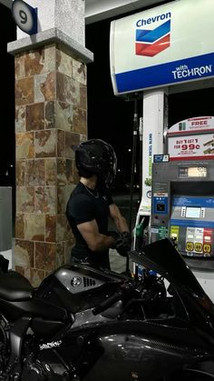 a man in black shirt and helmet standing next to motorcycle at gas pump with chevron sign