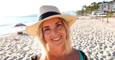 a woman with blonde hair wearing a hat and smiling at the camera on a beach