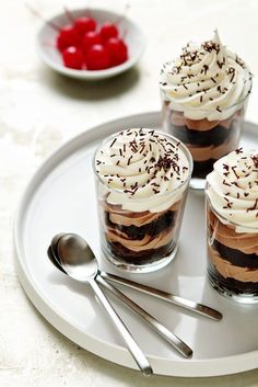 three desserts on a plate with spoons and cherries in the bowl behind them