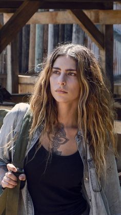 a woman with long hair and tattoos standing in front of a wooden structure, looking at the camera