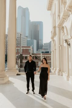 an engaged couple holding hands and walking in front of the columns of a large building