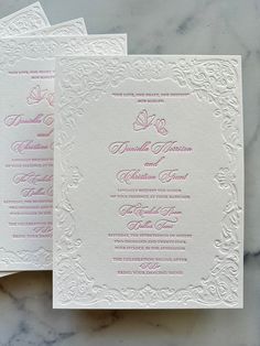 two white and pink wedding cards sitting on top of a marble countertop next to each other