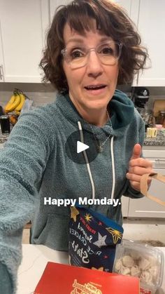 a woman in glasses is holding a box of cookies and pointing to the camera with her thumb