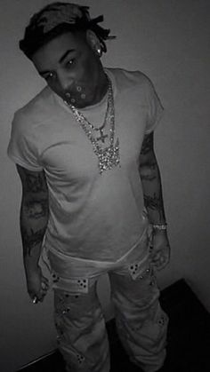 a man with tattoos and piercings standing in front of a wall wearing white pants