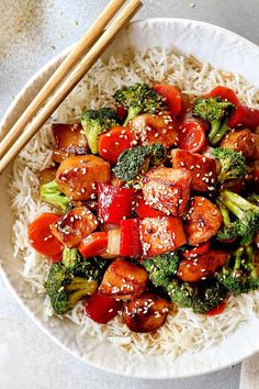 a white bowl filled with broccoli, chicken and rice next to chopsticks