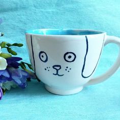 a coffee cup with a cat's face painted on it next to purple flowers