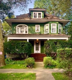 a green house with white trim on the front door and two story windows, surrounded by greenery