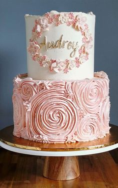 a three tiered cake with pink icing and flowers on top, sitting on a wooden stand