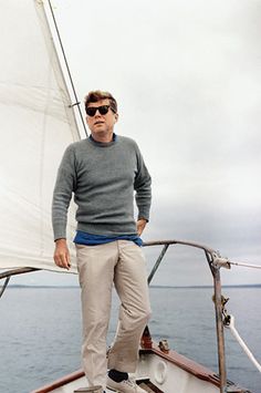 a man standing next to a sailboat on the water with his hands in his pockets