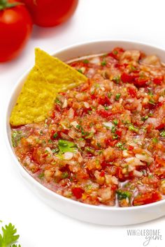 a white bowl filled with salsa next to two tomatoes and a tortilla chip