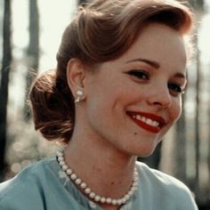 a woman in a blue dress smiling and wearing pearls on her necklace, with trees in the background