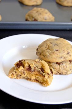 two chocolate chip cookies on a white plate next to a baking pan with muffins in the background