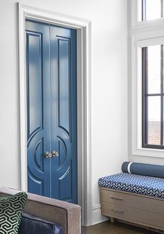 a blue door in a white room next to a bench and window with pillows on it