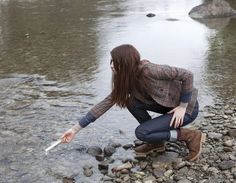 a woman crouches down to pick up rocks from the water's edge while holding an object in her hand