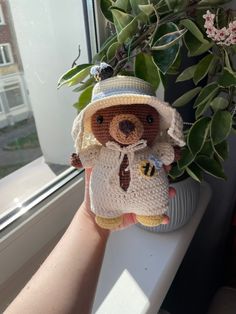 a hand holding a teddy bear next to a potted plant on a window sill