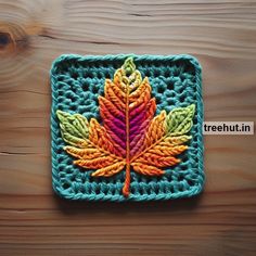 a crocheted square with a colorful leaf on the front and bottom, sitting on a wooden surface