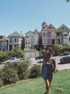 a woman in overalls is walking on the grass near some houses with cars parked outside