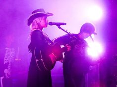 two people standing on stage with guitars and microphones in front of purple light behind them