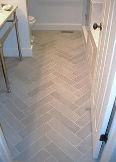 a bathroom with white walls and grey herringbone tile flooring, along with an open door leading to the toilet