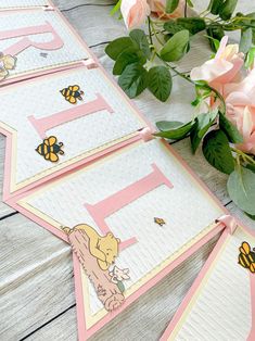 three pink and white cards with bees on them sitting next to some flowers, one has the letter e