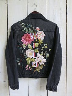 an embroidered denim jacket hanging on a white wooden wall with flowers and letters painted on it