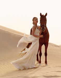 a woman in a white dress standing next to a brown horse on top of a sandy hill