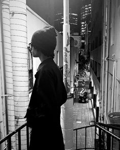 black and white photograph of woman standing on balcony overlooking city street at night with traffic passing by