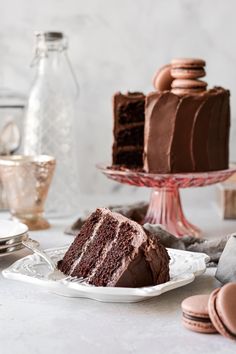 a slice of chocolate cake on a white plate with cookies and milk in the background