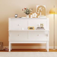 a white dresser sitting on top of a wooden floor next to a lamp and pictures