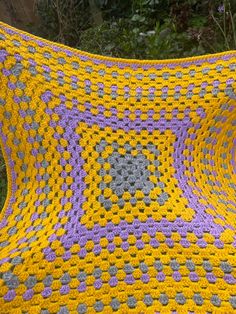 a crocheted blanket that is yellow and purple with flowers on the outside, in front of some bushes