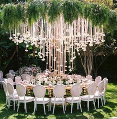 an outdoor dining table with white chairs and chandelier hanging from the ceiling is surrounded by lush greenery