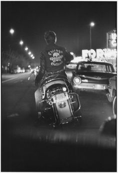 black and white photograph of man on motorcycle in street at night with cars behind him