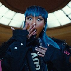 a woman with blue hair and piercings covers her mouth while standing in front of a round window