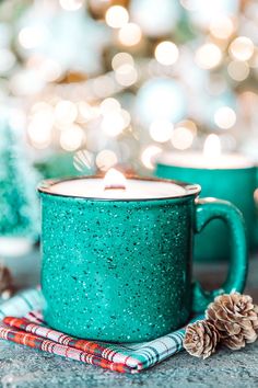 two teal mugs sitting next to each other with pine cones on the table