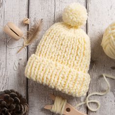 a white knitted hat next to a pair of scissors and some pine cones on a wooden table