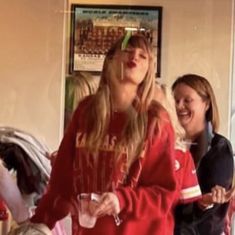 two young women are laughing and drinking in a room full of other people, one is wearing a red sweatshirt