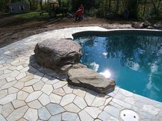 a stone patio around a pool with a large rock in the middle and a man walking by