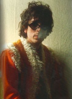 a man wearing sunglasses and a knitted vest standing in front of a white wall